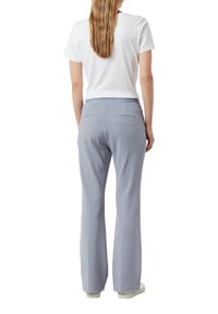 Osoba z długimi włosami ubrana w białą koszulę z krótkim rękawem i jasnoniebieskie, szerokie spodnie, stojąca tyłem na białym tle.