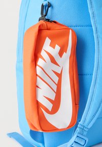 Φωτεινό πορτοκαλί τσάντα Nike με μεγάλο λευκό λογότυπο προσαρτημένο στο πλευρικό λουράκι μιας ανοιχτόχρωμης γαλάζιας τσάντας πλάτης.