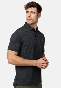 Polo noir chiné avec un col classique, patte de boutonnage à trois boutons et logo orange contrastant sur la poitrine gauche. Manches courtes.