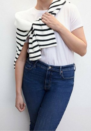 Camiseta blanca, jeans oscuras y un suéter a rayas blanco y negro colgado sobre el hombro. Primer plano del torso y el brazo.