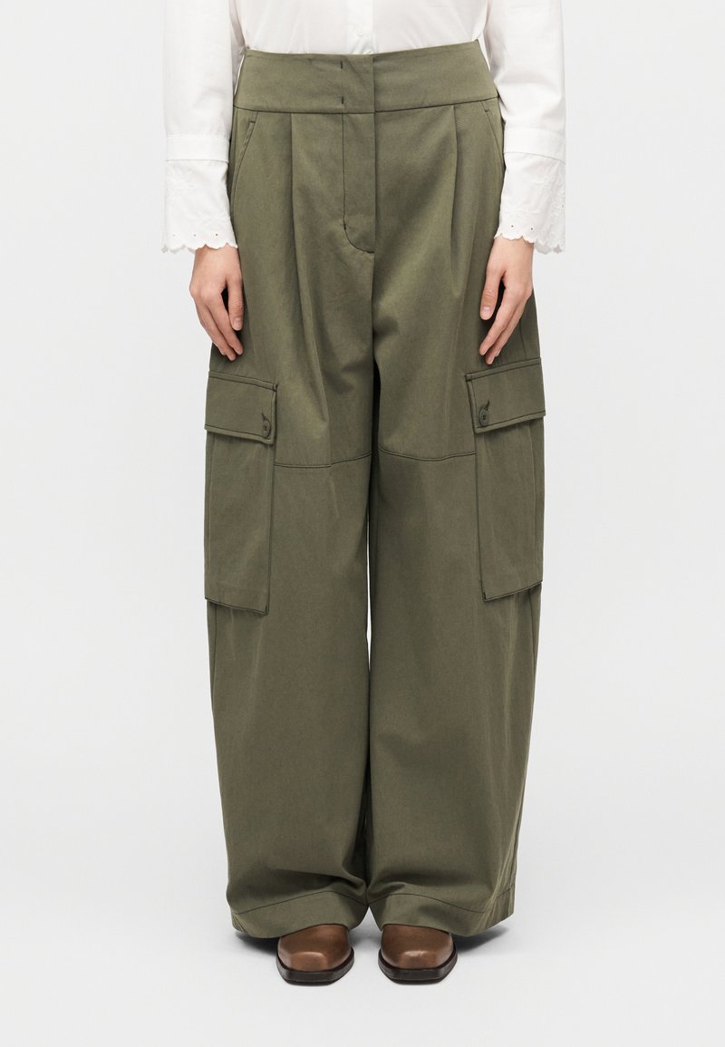 Πράσινο παντελόνι εργασίας με φαρδιά γραμμή, εμπ ρός πιέτες και μεγάλες πλαϊνές τσέπες, κατασκευασμένο από ανθεκτικό ύφασμα. Καφέ παπούτσια φαίνονται από κάτω.