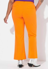 Pantaloni arancioni svasati con una texture liscia, dotati di due tasche posteriori. Abbinati a stivali alla caviglia metallici argento e un design minimalista.
