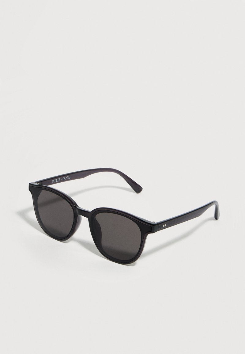 Μαύρα γυαλιά ηλίου με πλαστική σκελετό και σκουρόχρωμους φακούς, με ελαφρώς στρογγυλεμένο τετράγωνο σχήμα, που βρίσκονται πάνω σε λευκή επιφάνεια.