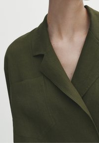 Giacca in lino verde oliva con colletto a incasso, tasca frontale e vestibilità rilassata. Tessuto liscio e design minimalista.