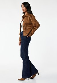Bruine suede jasje met klepzakken, knoopdetails; gecombineerd met donkerblauwe flared jeans en bruine hoge laarzen.