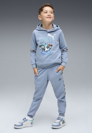 Junger Junge steht mit den Händen in den Taschen, trägt einen hellblauen Puma-Hoodie, passende Sweatpants und blau-weiße Sneaker vor einem grauen Hintergrund.