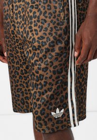 Brown leopard print shorts featuring black accents, white side stripes, and a small white logo at the bottom left. Made from a smooth fabric.