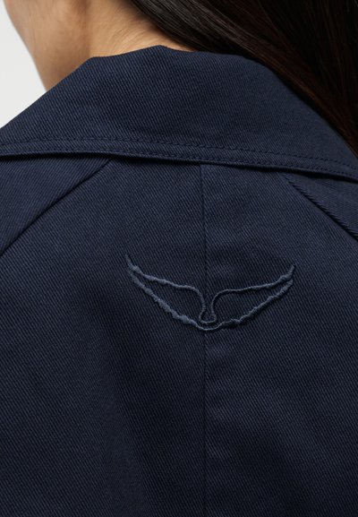 Μπροστινή όψη μπλε ναυτικού σακακιού με γιακά και σκούρο κεντητό λογότυπο σε σχήμα φτερού στο κέντρο κάτω από τη γραμμή του λαιμού, φορεμένο από άτομο με μακριά σκούρα μαλλιά.