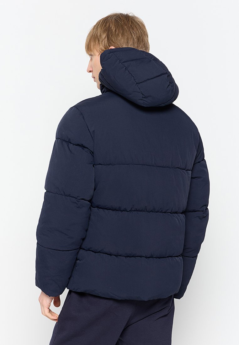 Veste en duvet navy avec capuche, présentant une silhouette boxy et un design matelassé. Fabriquée en matériau isolant avec des détails de coutures visibles.