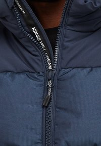 Tmavě modrá prošívaná bunda s vysokým límcem, výrazným zipem a texturovanou tkaninou, zdobená názvem značky na vnitřní podšívce.