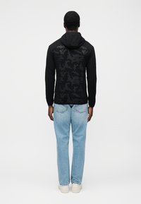 Μαύρο φούτερ με κουκούλα και υφασμένο μοτίβο παραλλαγής στην πλάτη, συνδυασμένο με ανοιχτό μπλε τζιν και λευκά παπούτσια τύπου αθλητικά.