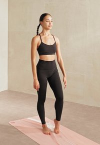 Musta urheilbra ja korkeavyötäröiset legginsit, henkilö seisoo paljain jaloin vaaleanpunaisella jooga-alustalla, jossa on sileä, matta pinta.