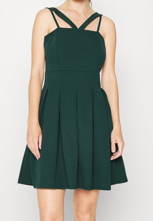 Robe sans manches vert foncé avec de larges et fines bretelles doubles et une jupe plissée portée par une personne debout.