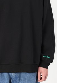 Sweatshirt preto com punhos e bainha canelados, apresentando um logo "adidas" bordado em verde na manga esquerda. Textura de tecido suave.