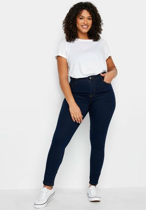 Femme aux cheveux bouclés portant un t-shirt blanc, un jean taille haute bleu foncé et des baskets blanches, debout avec une main dans la poche, souriante.