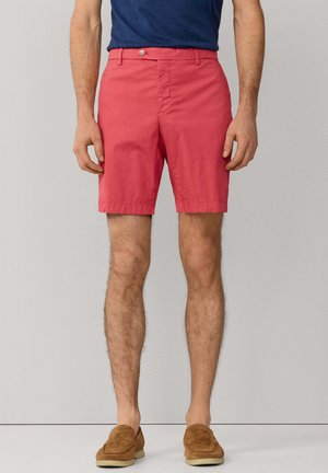 Man draagt koraalkleurige korte broek tot halverwege de dij, marineblauw overhemd ingestopt en bruine suède instapschoenen, staand tegen een lichtgrijze achtergrond.