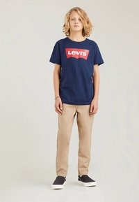 Tmavě modré bavlněné tričko s velkým červeným logem Levi's, zkombinované s béžovými kalhotami a černými nazouvacími botami. Jednoduchý design, volný střih.