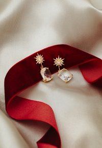 Gold-tone earrings with star-shaped accents and large clear gemstones, displayed on a red ribbon against a soft beige fabric background.