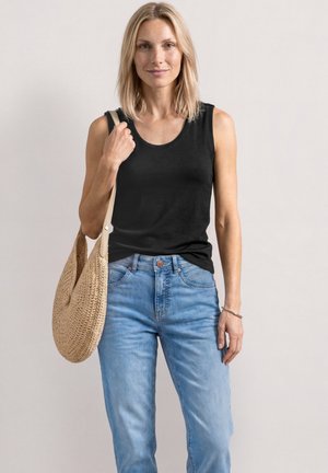 Blonde Frau trägt ärmelloses schwarzes Oberteil und hellblaue Jeans, hält eine große geflochtene Schultertasche, steht vor einfachem Hintergrund.