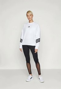 Sweatshirt branco com realces de três riscas pretas, combinado com leggings pretas com painéis em rede. Modelo de pé em sapatilhas atléticas brancas.