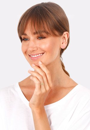 Goldring mit filigranem Design, getragen an einer Hand vor einem hellen Hintergrund. Die Trägerin hat mittelbraunes Haar und lächelt. Weißes Oberteil.