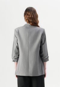 Blazer gris avec une coupe cintrée, manches retroussées et texture lisse. Comprend un col cranté et un détail de couture au dos.