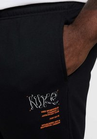 Pantalones de chándal de algodón negros con un logo de Nike bordado, detalles en naranja y bolsillos laterales. Ajuste suave y relajado con una textura lisa.