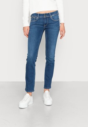 Dark blue denim jeans with a slim fit, featuring five pockets and a light wash effect. Paired with white platform sneakers.
