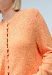 Cardigan en tricot orange avec col rond et fermeture à boutons. Tissu texturé avec des boutons proéminents pour la fermeture et le détail.