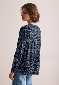 Långärmad marinblå blus med ett ljusblått abstrakt mönster, avslappnad passform och en veckdetalj bak, i kombination med ljusblå jeans.