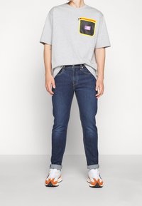 Šedé tričko s krátkým rukávem a síťovou kapsou s jasně žlutým lemem. Spárované s tmavě modrými džíny a bílými teniskami s oranžovými akcenty.