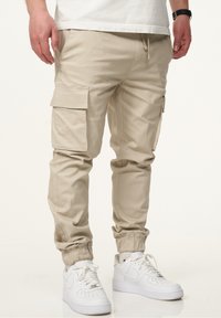 Beige Cargo-Hosen aus einem Baumwollmischgewebe, ausgestattet mit Seitentaschen, elastischen Bündchen und einem Kordelzugbund, kombiniert mit weißen Turnschuhen.