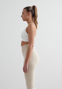 Witte sportbeha en beige hoge taille legging gedragen door een persoon gezien vanaf de zijkant, die een gladde, strakke vorm en stoftextuur laat zien.