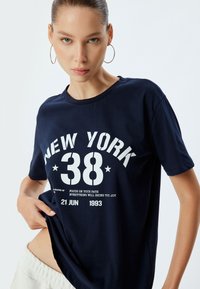 Camiseta de algodón azul marino con gráficos blancos llamativos: "NUEVA YORK 38," con texto adicional y cuello redondo. Mangas cortas, corte relajado.