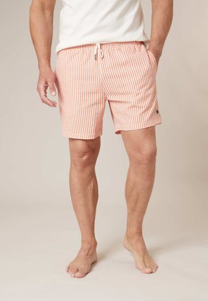 Man die oranje-wit gestreepte korte broek met verticale strepen en een witte trekkoord draagt, op blote voeten staand tegen een effen achtergrond.