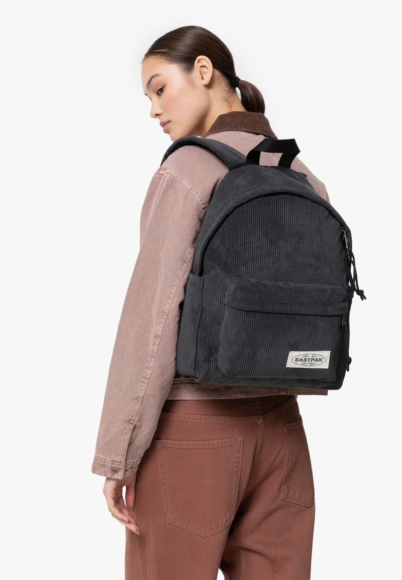 Zaino in corduroy nero con tasca frontale e patch con logo, indossato con una giacca rosa chiaro e pantaloni marroni, dotato di spallacci regolabili.