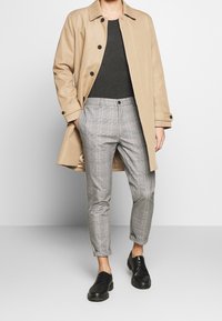 Beige trench coat over a gray top, plaid gray trousers with rolled cuffs, and black lace-up shoes. Smooth fabric with button details.