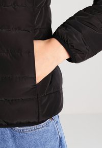 Detail einer schwarzen Daunenjacke, die eine Hand in einer Seitentasche zeigt, mit gestepptem Muster und elastischen Bündchen sowie einem glatten Finish.