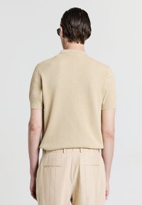 Beige gestricktes Polo-Shirt mit kurzen Ärmeln, strukturiertem Muster und geripptem Kragen, kombiniert mit beigen gestreiften Hosen.