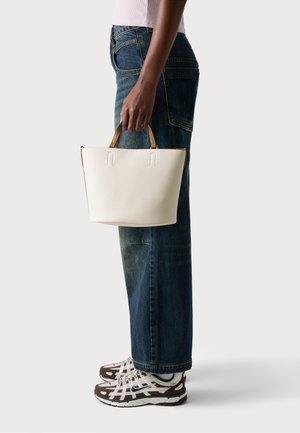 Sac à main texturé blanc avec des poignées dorées, forme cylindrique et sans logos visibles, associé à un jean foncé et des baskets.