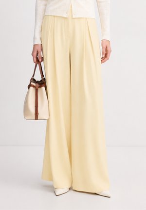 Persona che indossa pantaloni giallo pallido a gamba larga, scarpe bianche appuntite, che tiene una borsa crema e marrone con manici rotondi.