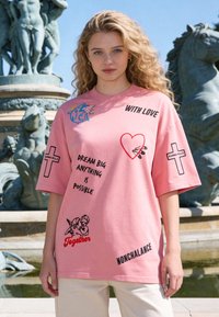 Jeune femme aux cheveux blonds bouclés portant une chemise rose avec des phrases, des dessins et des croix, debout devant une sculpture de fontaine.
