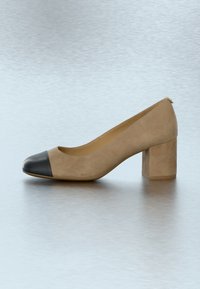 Beige ruskindsstilet med en rundet tå, der har en sort læderkappe foran og en klodset hæl. Glat tekstur med minimale designdetaljer.