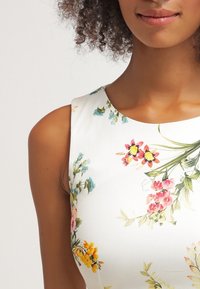 Vestido de estampado floral con tejido blanco ajustado, presenta vibrantes flores en tonos rosa, amarillo y azul, escote redondo y diseño sin mangas.