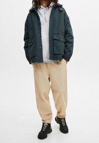 Person som bär en mörkgrön jacka med stora fickor, ljusgrå hujacka, beige manchestertygsbyxor och svarta vandringsstövlar, stående mot en vit bakgrund.