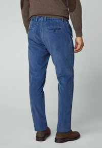 Homme portant un pantalon en velours côtelé bleu, des chaussures en daim marron et un pull marron avec des coudières, debout devant un fond uni.