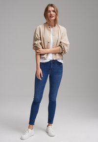 Beige bomber jacket with gathered sleeves, worn over a white shirt, paired with dark blue skinny jeans and white sneakers. Simple design.