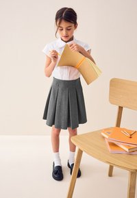 Jupe plissée grise, chemise blanche à col, mocassins noirs et chaussettes blanches. Tenant un dossier marron avec des pages colorées. Chaise en bois et carnets orange à proximité.