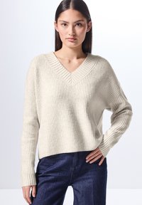 Maglione a righe color beige chiaro con scollo a V e maniche lunghe. Presenta una vestibilità comoda e un tessuto testurizzato. Abbinato a pantaloni in denim blu scuro.