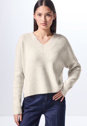 Helles beigefarbenes, geripptes Sweater mit V-Ausschnitt und langen Ärmeln. Hat eine lässige Passform und strukturierte Stoffqualität. Getragen mit dunkelblauen Jeanshosen.